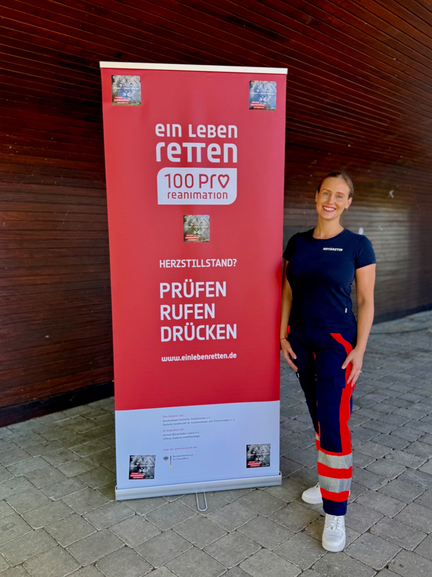 Anna Vormeyer in Notärztin-Kleidung steht neben dem roten Plakat „Ein Leben retten — 100 Pro Reanimation“ mit dem Motto Prüfen, Rufen, Drücken.